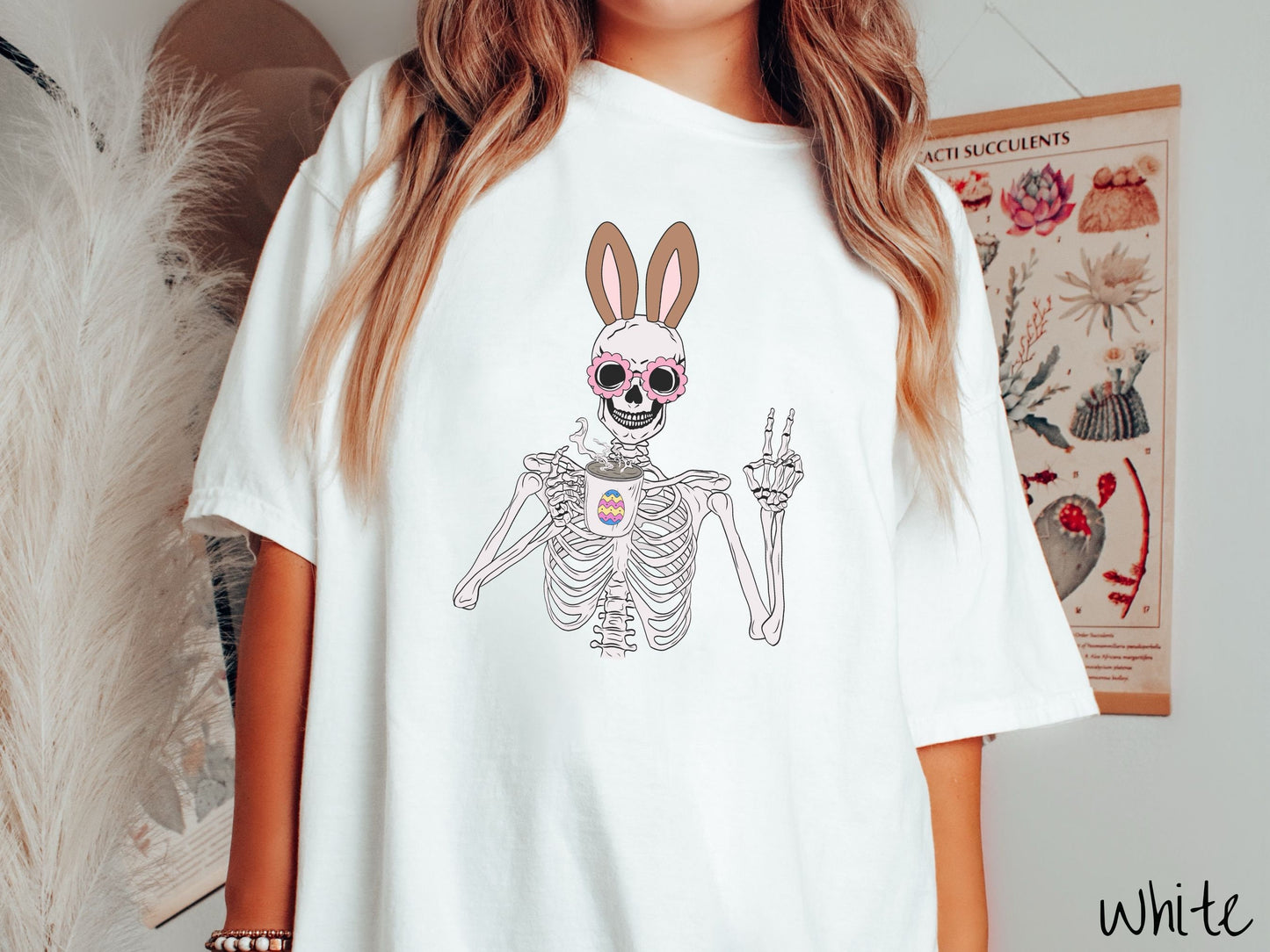 A woman wearing a cute, vintage white colored shirt with a pink-toned skeleton wearing pink glasses and brown bunny ears, holding a steamy cup of coffee while holding up a peace sign with the other hand. The white coffee cup has a colorful Easter egg