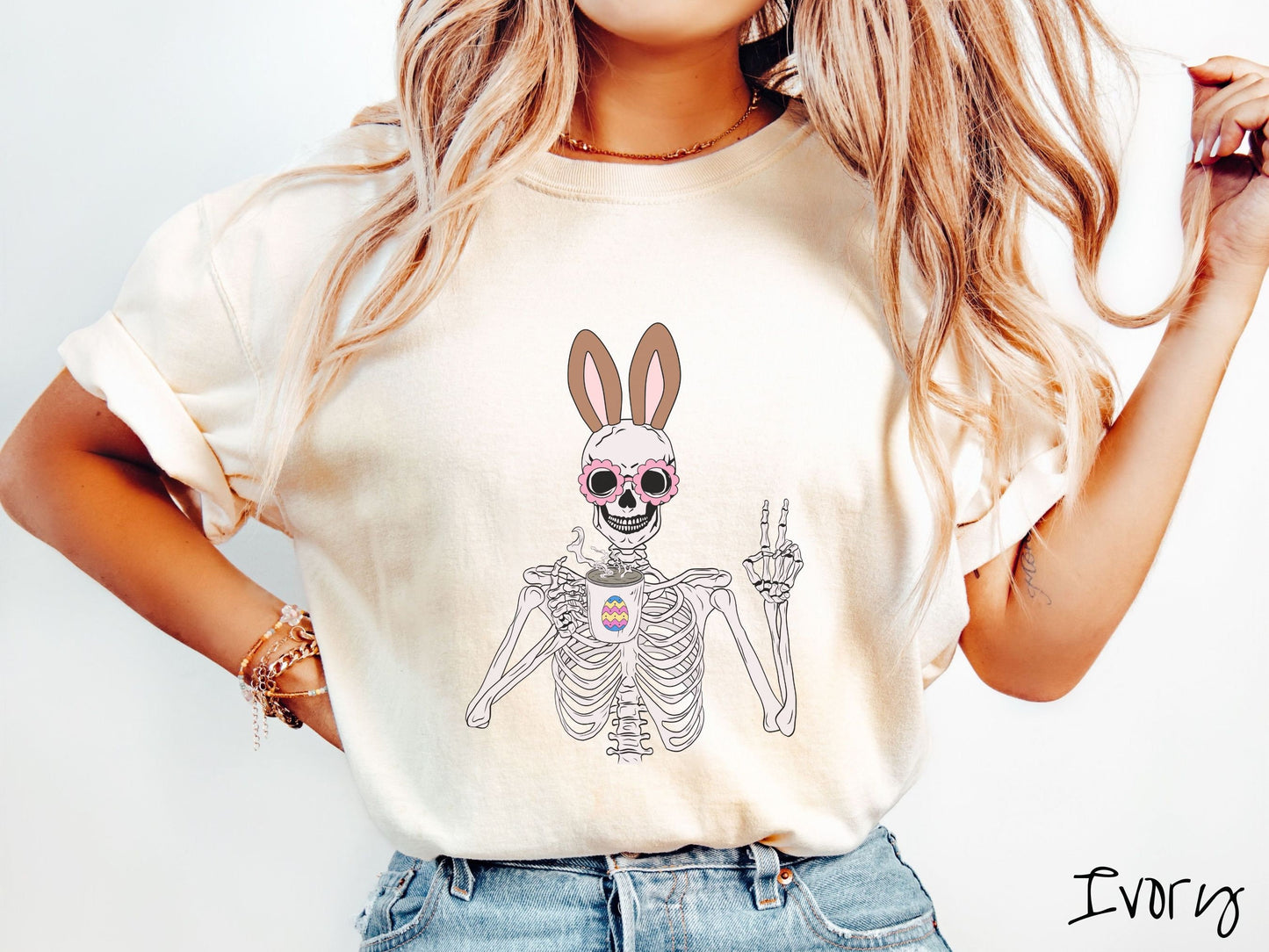 A woman wearing a cute, vintage ivory colored shirt with a pink-toned skeleton wearing pink glasses and brown bunny ears, holding a steamy cup of coffee while holding up a peace sign with the other hand. The white coffee cup has a colorful Easter egg