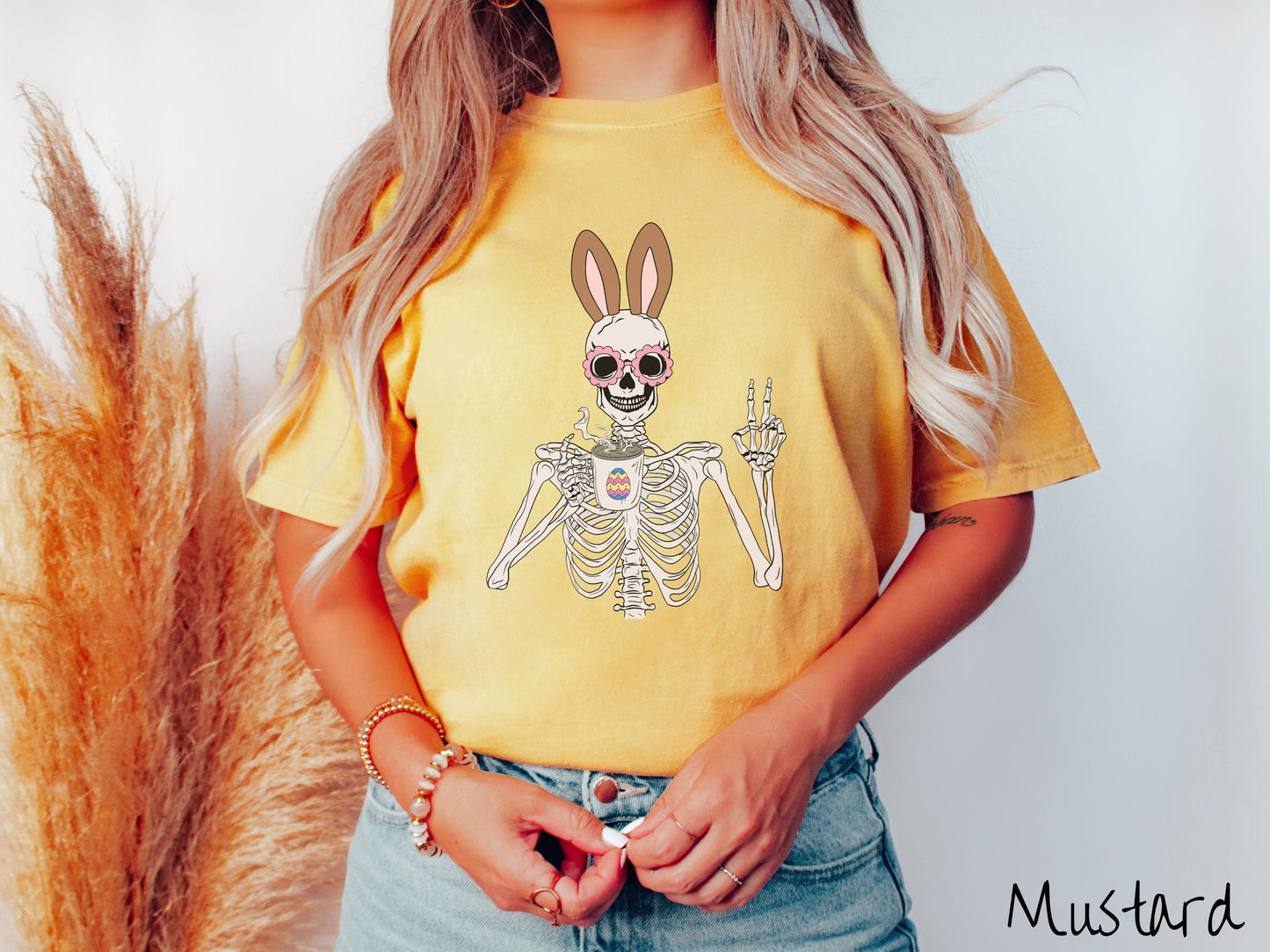 A woman wearing a cute, vintage mustard colored shirt with a pink-toned skeleton wearing pink glasses and brown bunny ears, holding a steamy cup of coffee and holding up a peace sign with the other hand. The white coffee cup has a colorful Easter egg