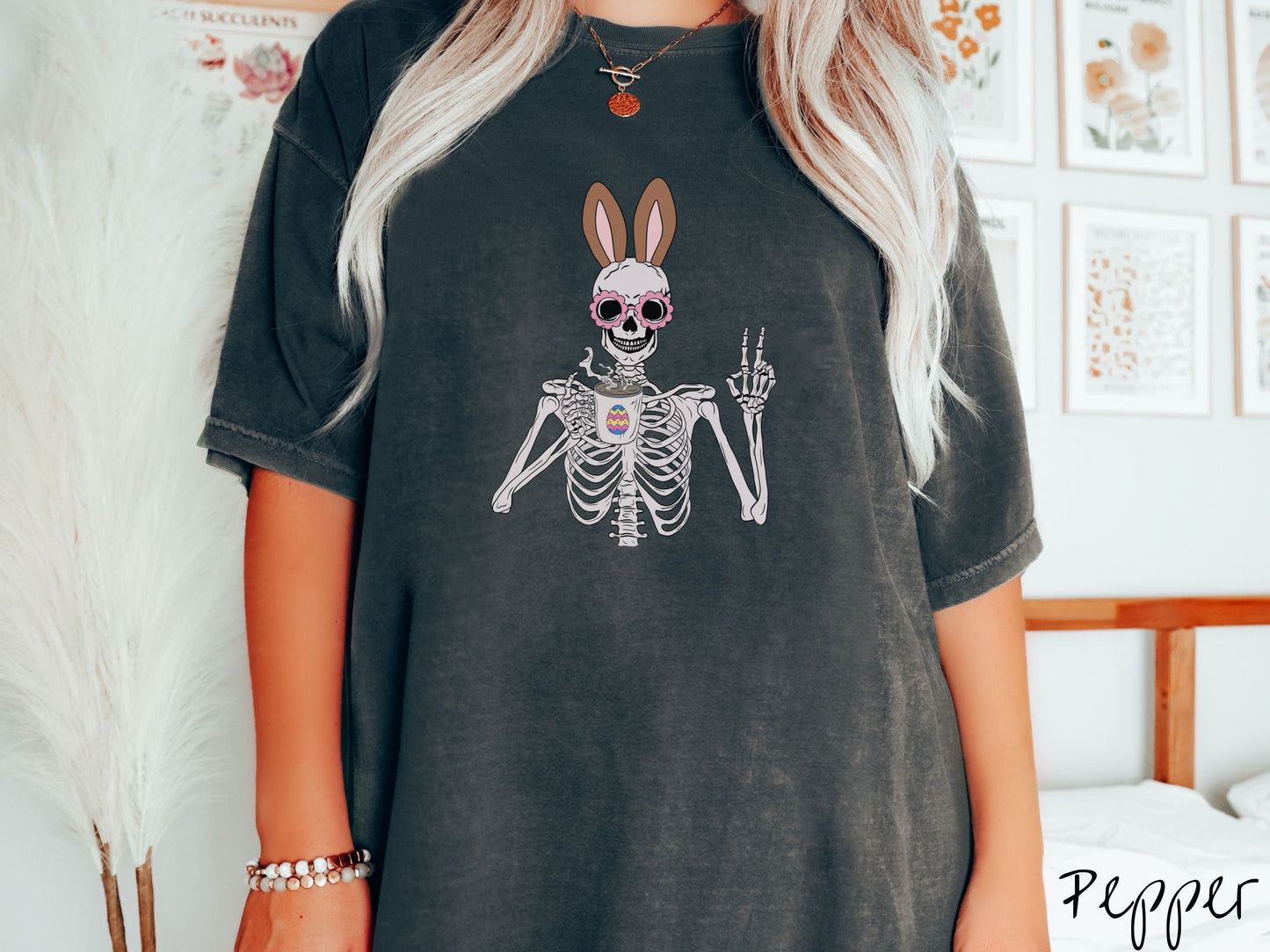 A woman wearing a cute, vintage pepper colored shirt with a pink-toned skeleton wearing pink glasses and brown bunny ears, holding a steamy cup of coffee and holding up a peace sign with the other hand. The white coffee cup has a colorful Easter egg