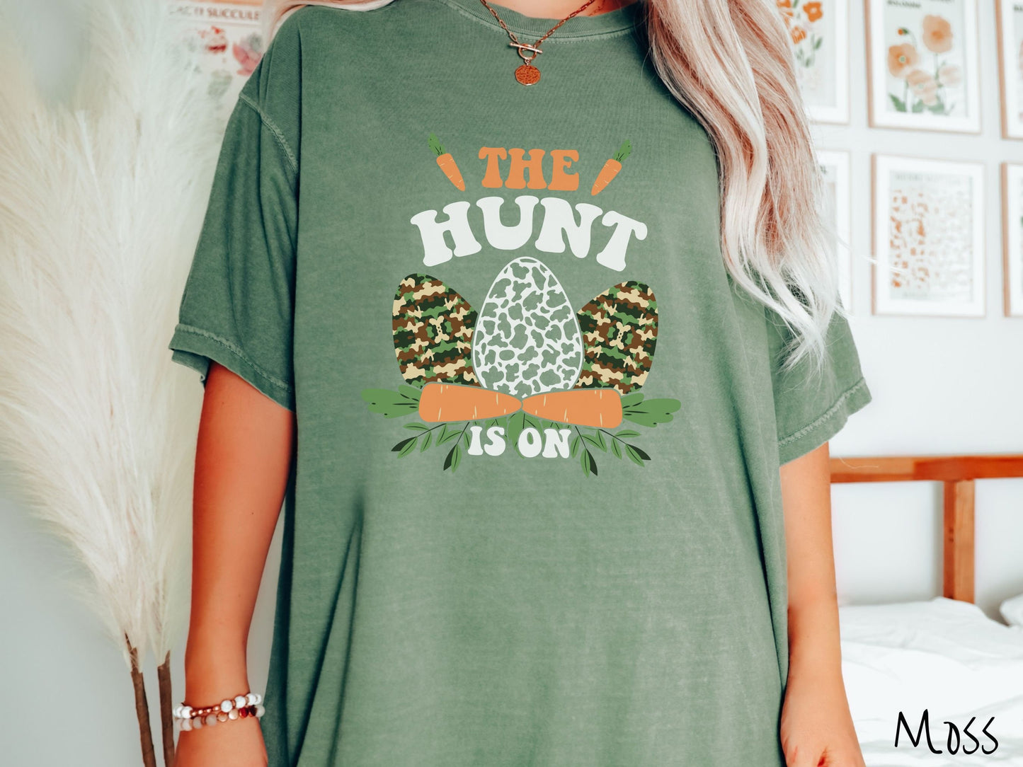 A woman wearing a cute, vintage moss colored shirt with the text The Hunt Is On in orange and white font, with three Easter eggs painted in woodland camo and black and white camo in front. Above and below the eggs are carrots with leafy ends.