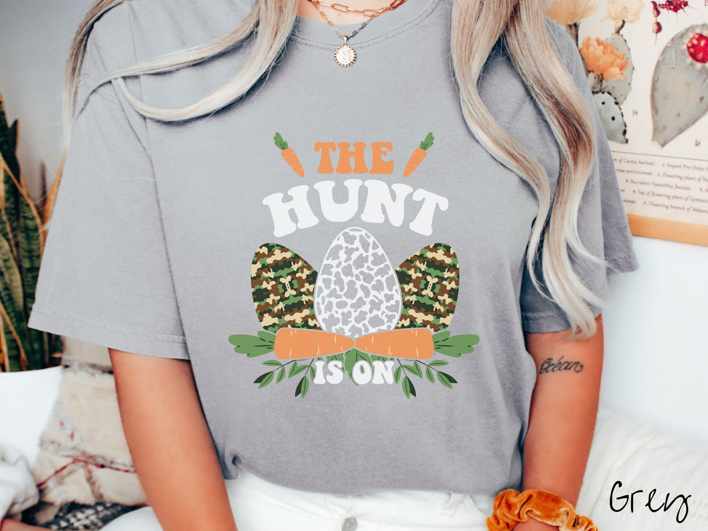 A woman wearing a cute, vintage grey colored shirt with the text The Hunt Is On in orange and white font, with three Easter eggs painted in woodland camo and black and white camo in front. Above and below the eggs are carrots with leafy ends.