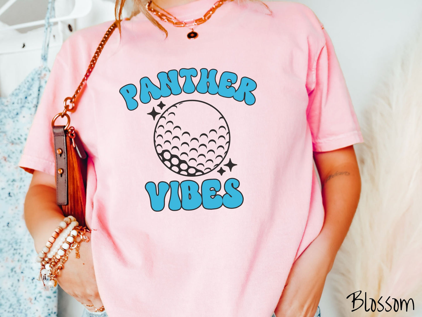 A woman wearing a cute blossom colored t-shirt with blue text saying Panther Vibes. There is a golf ball outline with black twinkling stars around it between the text.