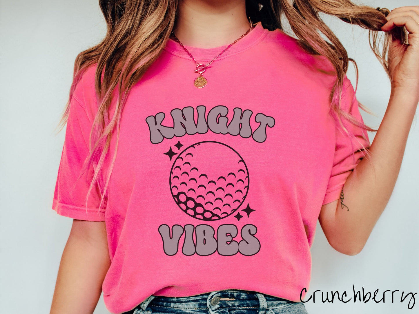 A woman wearing a cute crunchberry colored t-shirt with gray text saying Knight Vibes. There is a golf ball outline with black twinkling stars around it between the text.