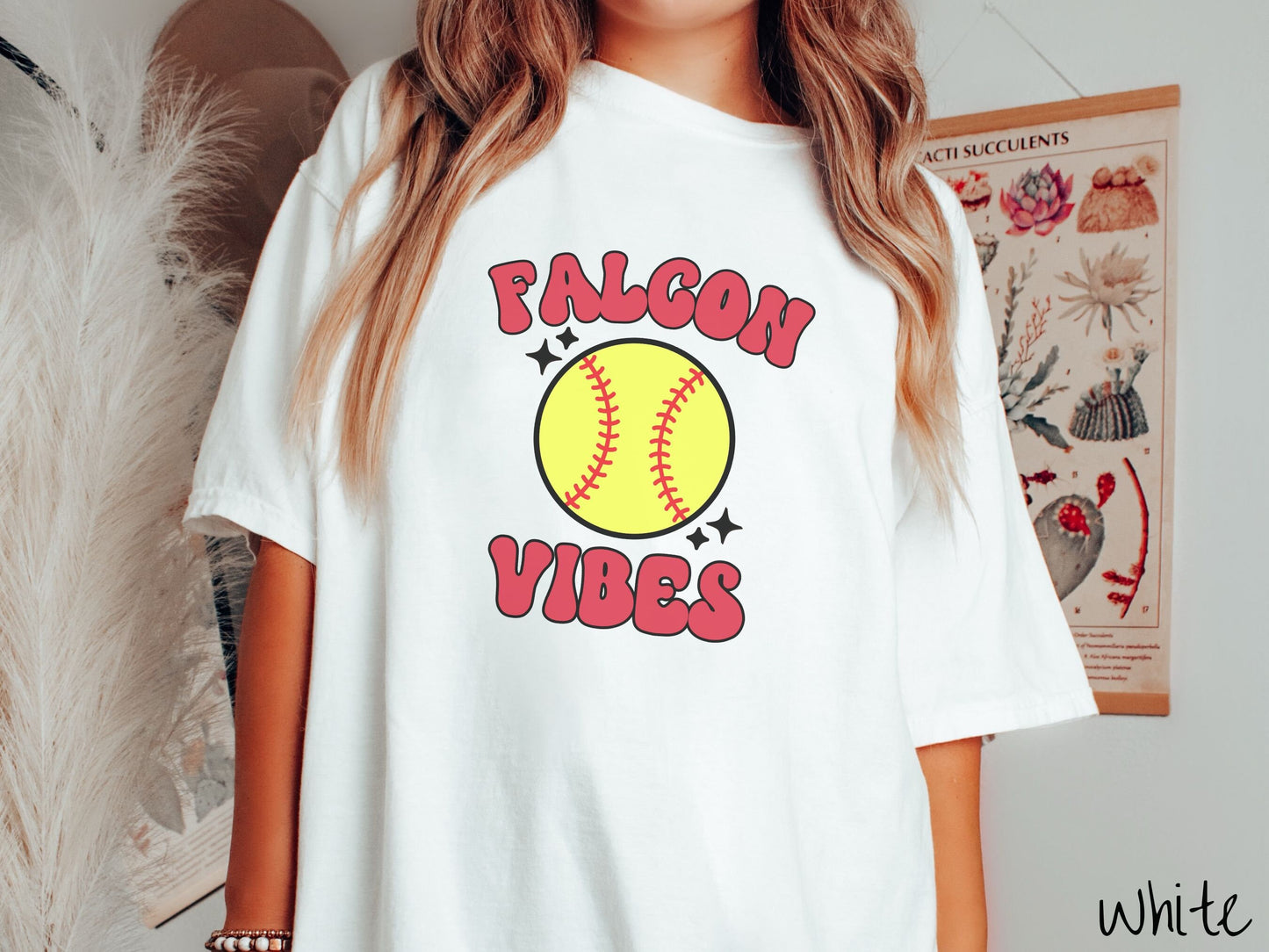 A woman wearing a cute white colored shirt with red text saying Falcon Vibes. There is a yellow baseball with red stitching with black twinkling stars around it between the text.