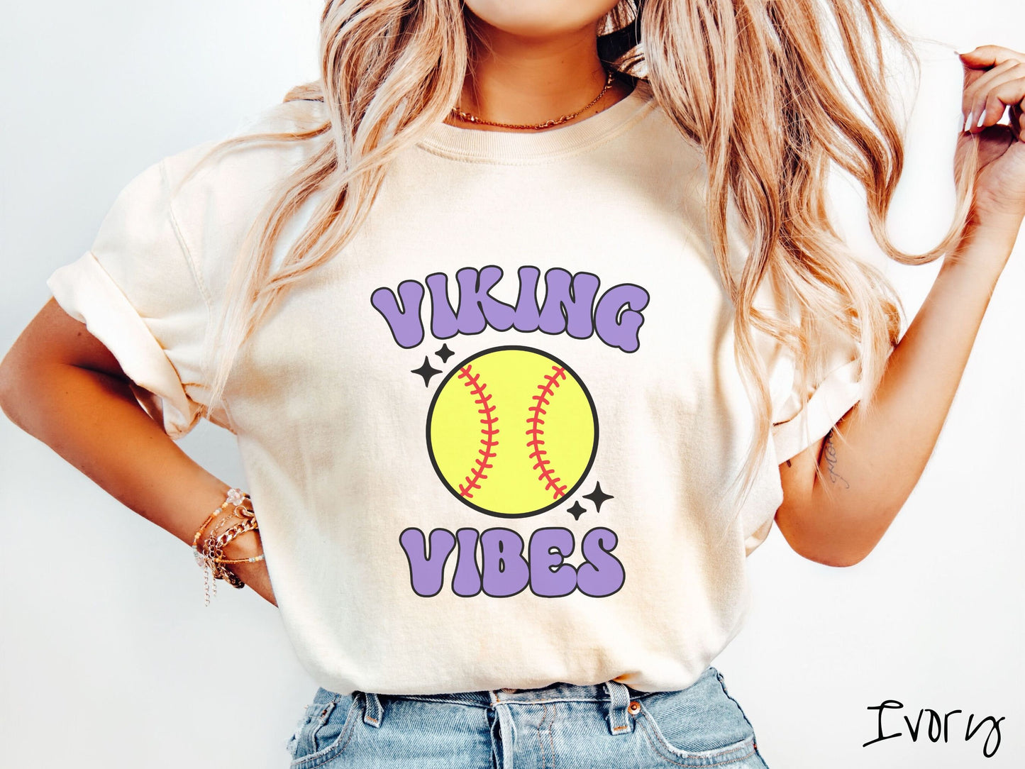 A woman wearing a cute ivory colored shirt with purple text saying Viking Vibes. There is a yellow baseball with red stitching with black twinkling stars around it between the text.