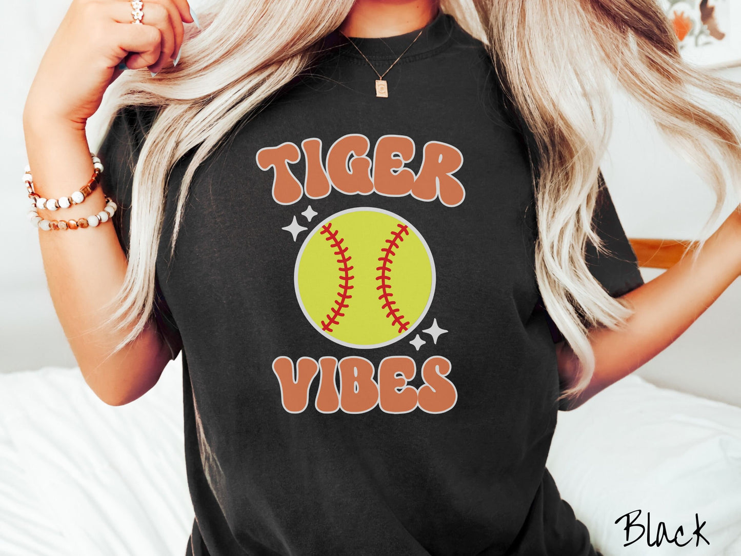 A woman wearing a cute black colored shirt with orange text saying Tiger Vibes. There is a yellow baseball with red stitching with black twinkling stars around it between the text.