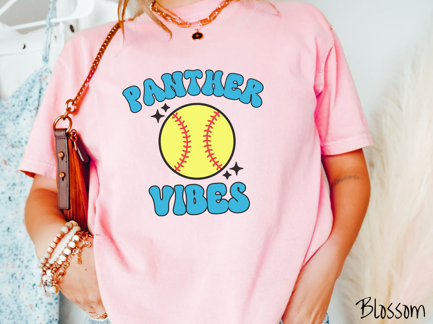 A woman wearing a cute blossom colored shirt with blue text saying Panther Vibes. There is a yellow baseball with red stitching with black twinkling stars around it between the text.