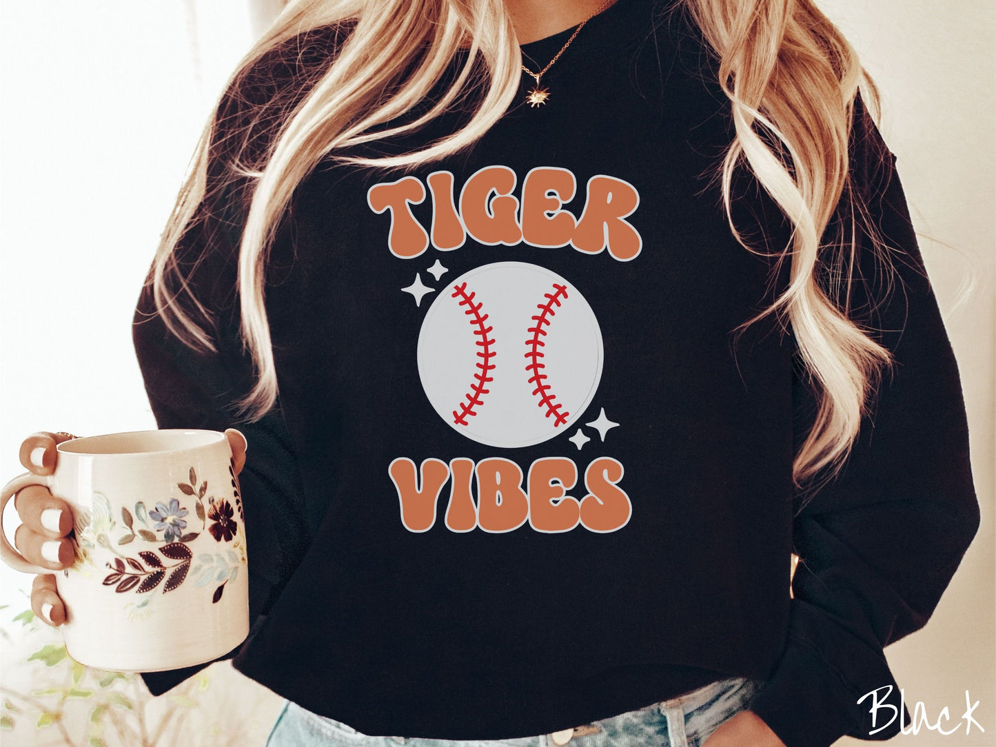 A woman wearing a cute black colored sweatshirt with orange text saying Tiger Vibes. There is a white baseball with red stitching between the text.