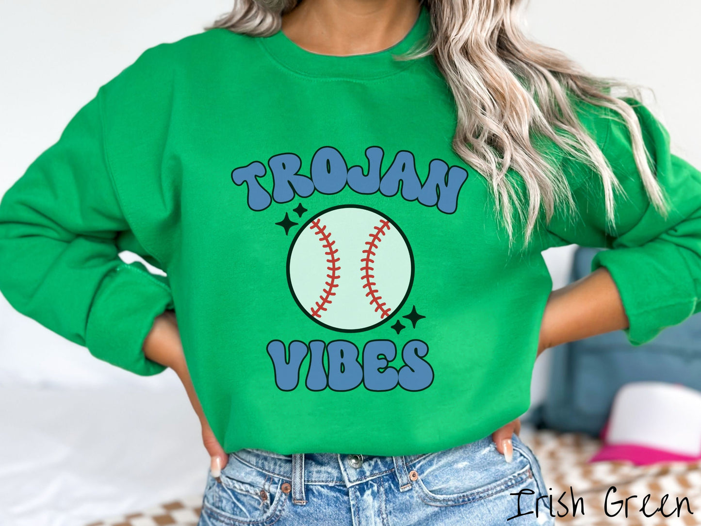 A woman wearing a cute Irish green colored sweatshirt with blue text saying Trojan Vibes. There is a white baseball with red stitching between the text.