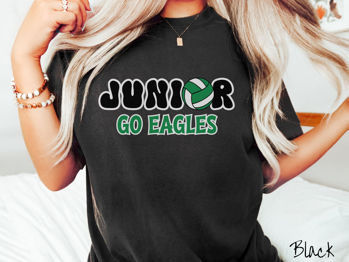 A woman wearing a cute black colored t-shirt with the word Junior in black and white font, the O in Junior is a green and white volleyball. Below Junior are the words in green and black font Go Eagles.