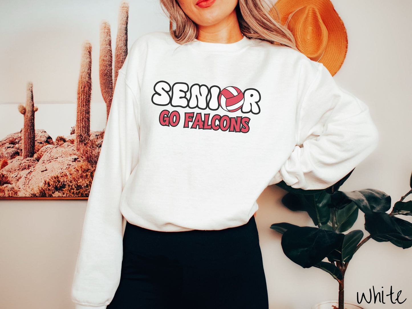 A woman wearing a cute white colored sweatshirt with the word Senior in black and white font, the O in Senior is a red and white volleyball. Below Senior are the words in red and black font Go Falcons.