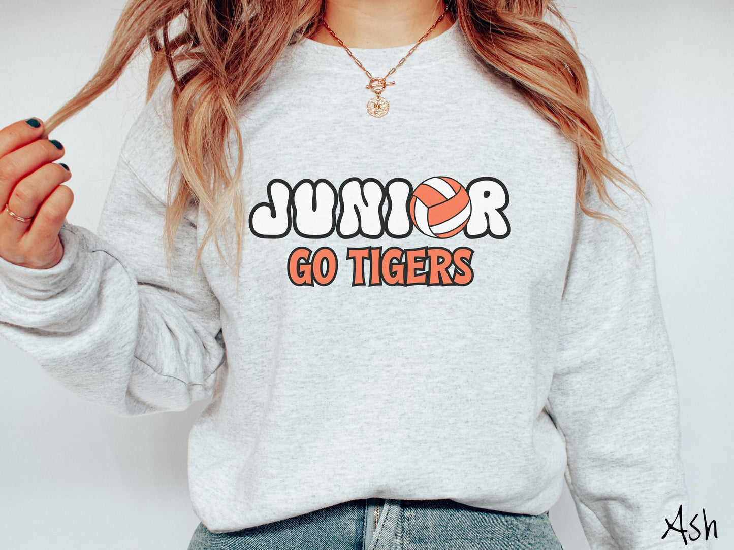 A woman wearing a cute ash colored sweatshirt with the word Junior in black and white font, the O in Junior is an orange and white volleyball. Below Junior are the words in orange and black font Go Tigers.