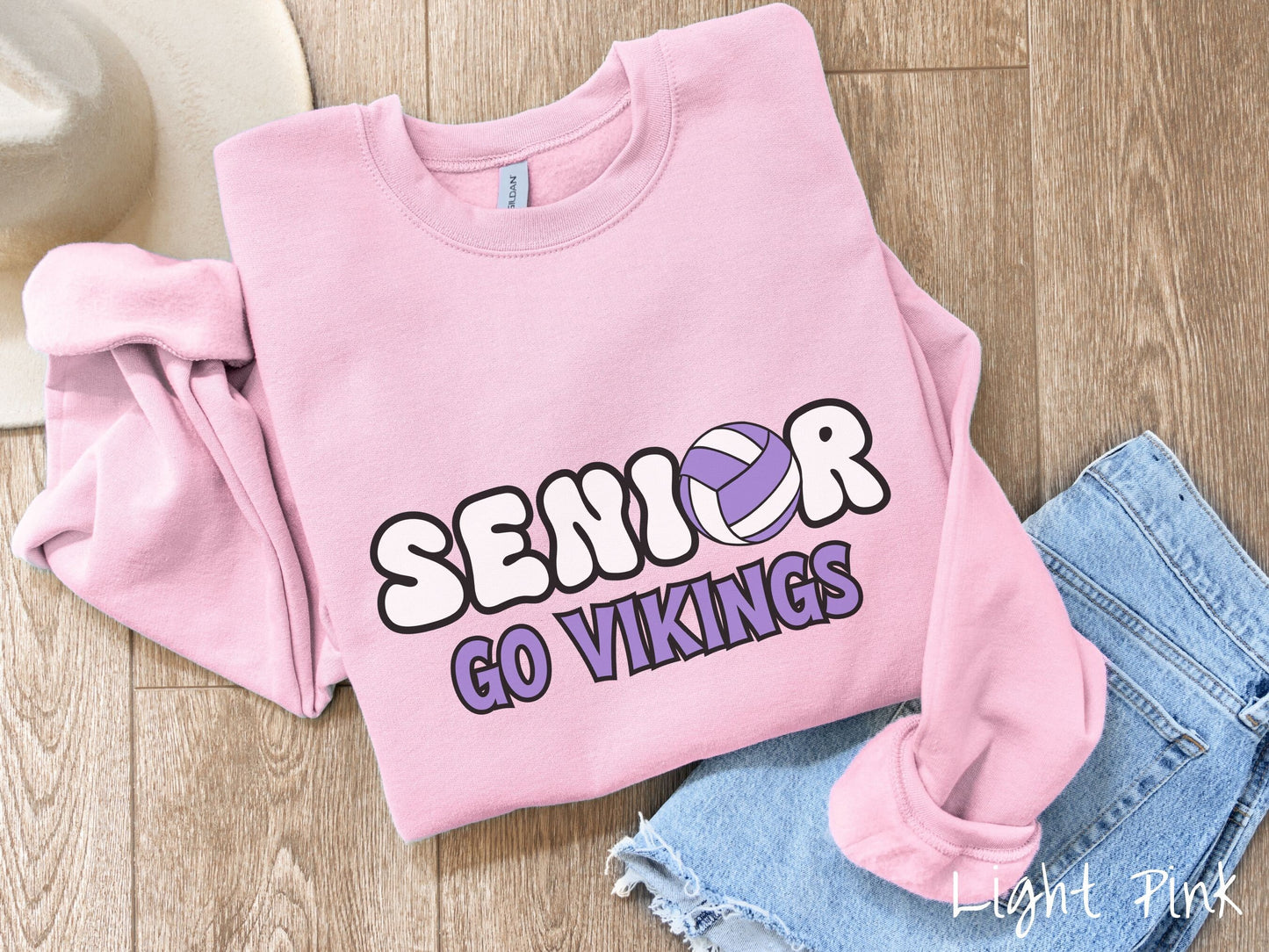 A woman wearing a cute light pink colored sweatshirt with the word Senior in black and white font, the O in Senior is a purple and white volleyball. Below Senior are the words in purple and black font Go Vikings.