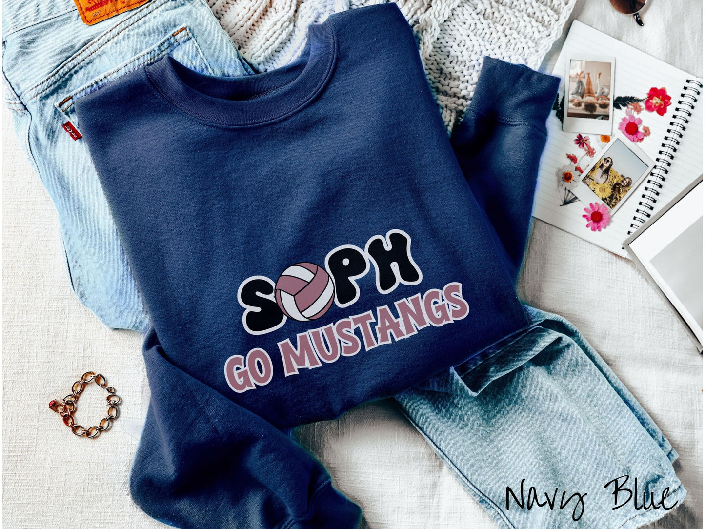 A woman wearing a cute navy blue colored sweatshirt with the word Soph in black and white font, the O in Soph is a purple and white volleyball. Below Soph are the words in purple and black font Go Mustangs.