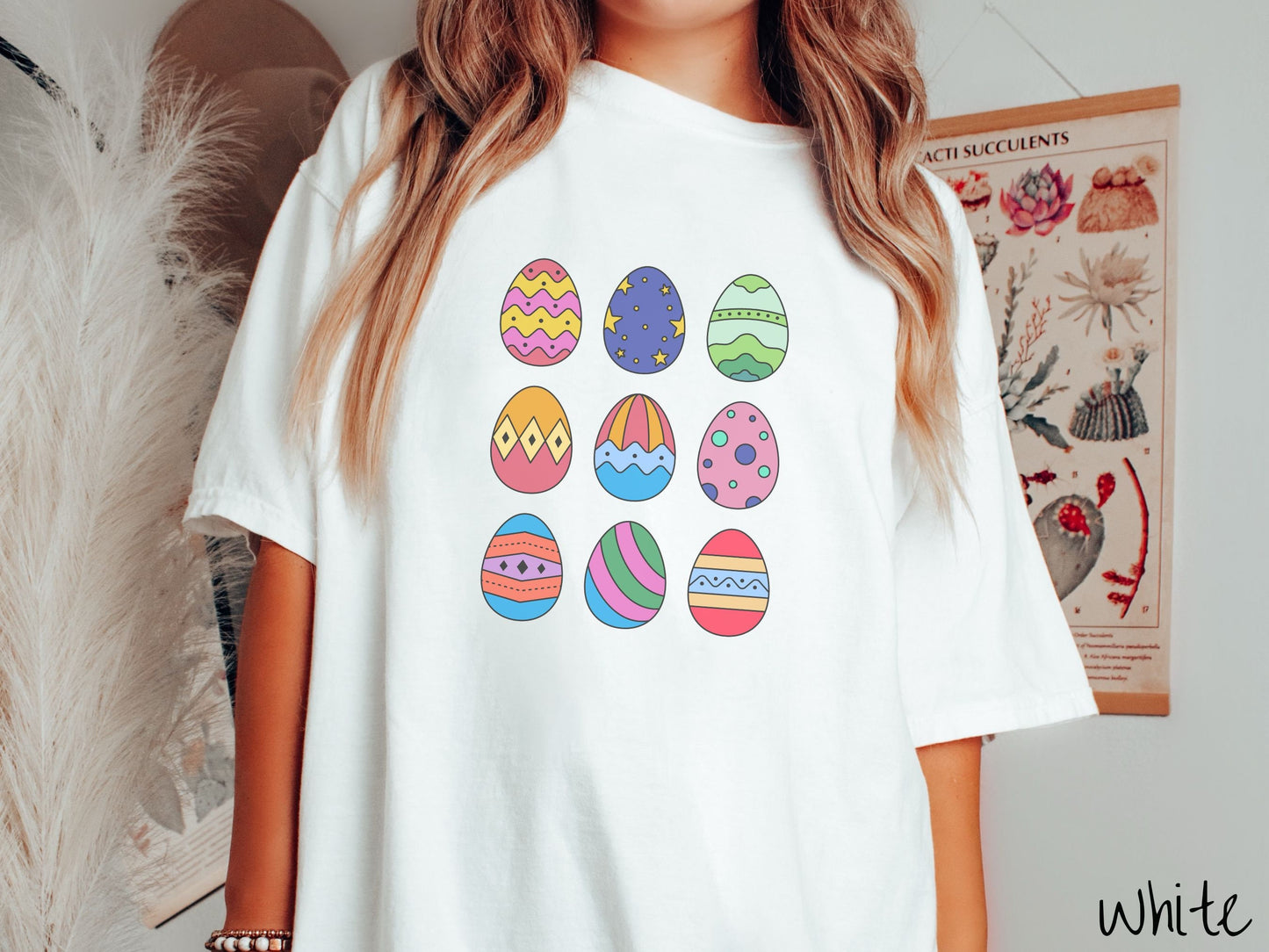 A woman wearing a cute, vintage white colored shirt with a three by three grid of colorful Easter eggs. One is pink, red, and yellow, another blue with stars, and another is different shades of green next to five more egg designs.