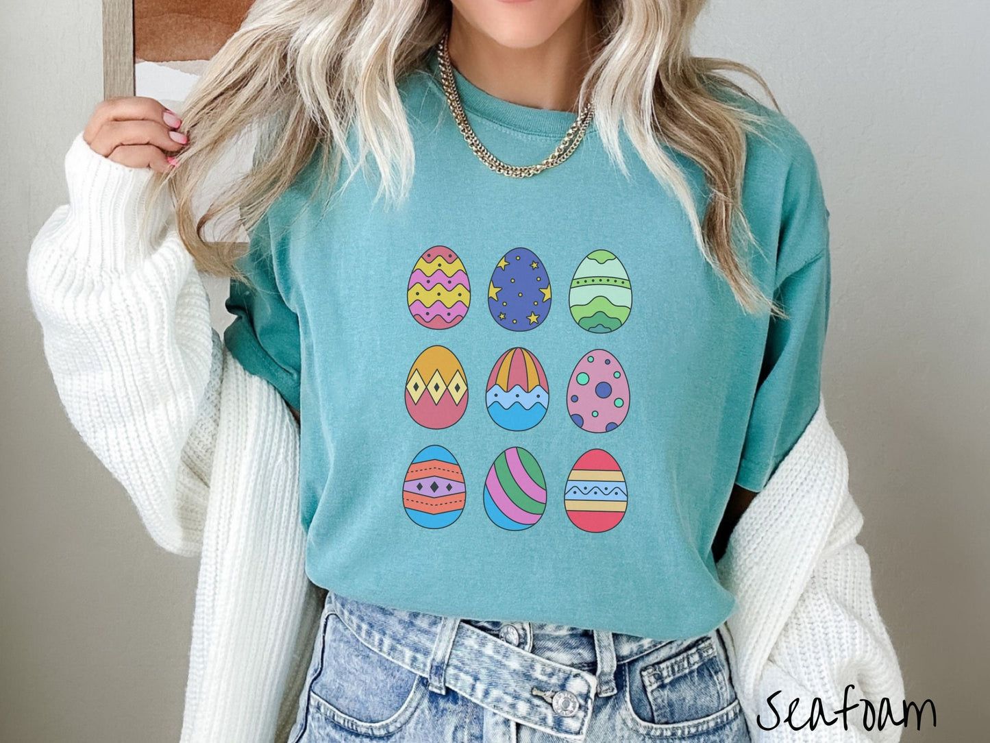 A woman wearing a cute, vintage seafoam colored shirt with a three by three grid of colorful Easter eggs. One is pink, red, and yellow, another blue with stars, and another is different shades of green next to five more egg designs.