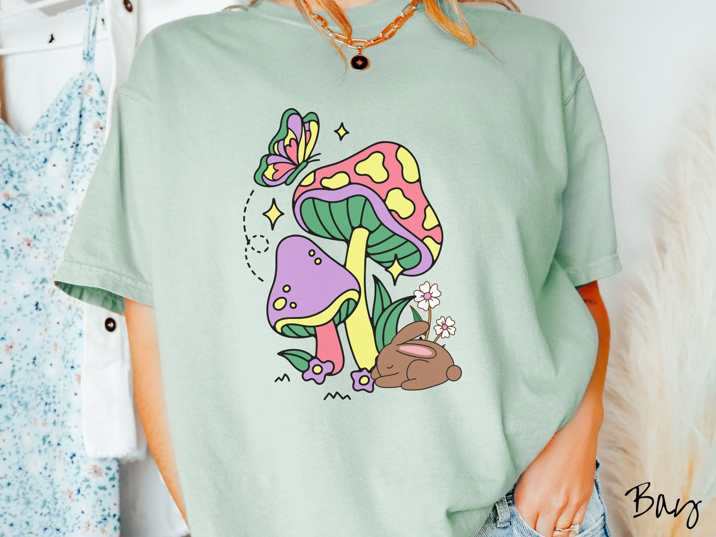 A woman wearing a cute, vintage bay colored shirt with a purple mushroom and a pink and yellow mushroom together with a brown bunny rabbit laying on the ground by purple and white flowers, and a colorful butterfly is fluttering above it all.