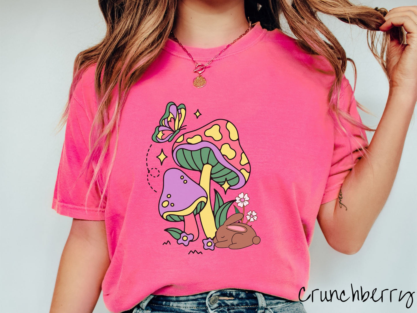 A woman wearing a cute, vintage crunchberry colored shirt with a purple mushroom and a pink and yellow mushroom together with a brown bunny rabbit laying on the ground by purple and white flowers, and a colorful butterfly is fluttering above it all.