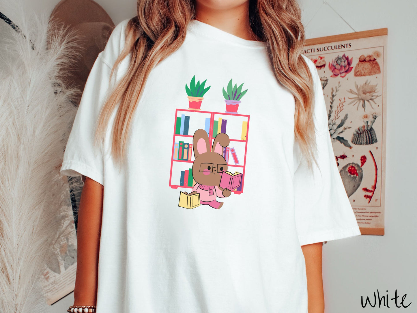 A woman wearing a cute, vintage white colored shirt with a pink-cheeked, brown bunny rabbit wearing glasses reading a pink book in one hand, and a yellow book in the other. In the background is a bookshelf with green plants on top.
