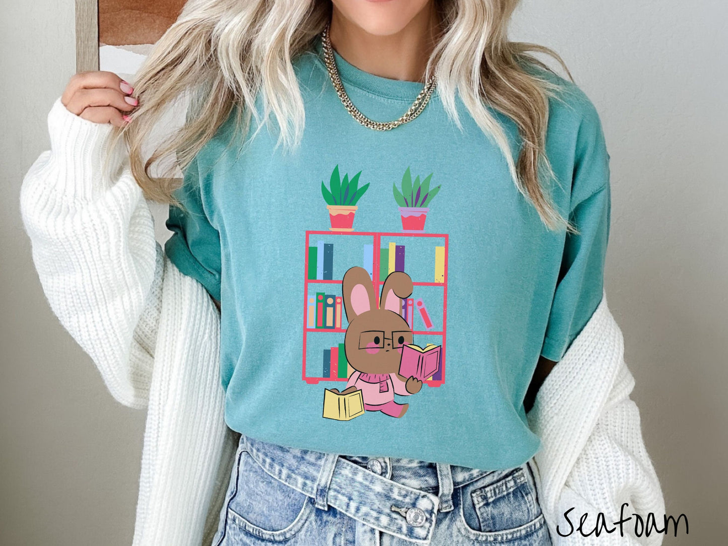 A woman wearing a cute, vintage seafoam colored shirt with a pink-cheeked, brown bunny rabbit wearing glasses reading a pink book in one hand, and a yellow book in the other. In the background is a bookshelf with green plants on top.
