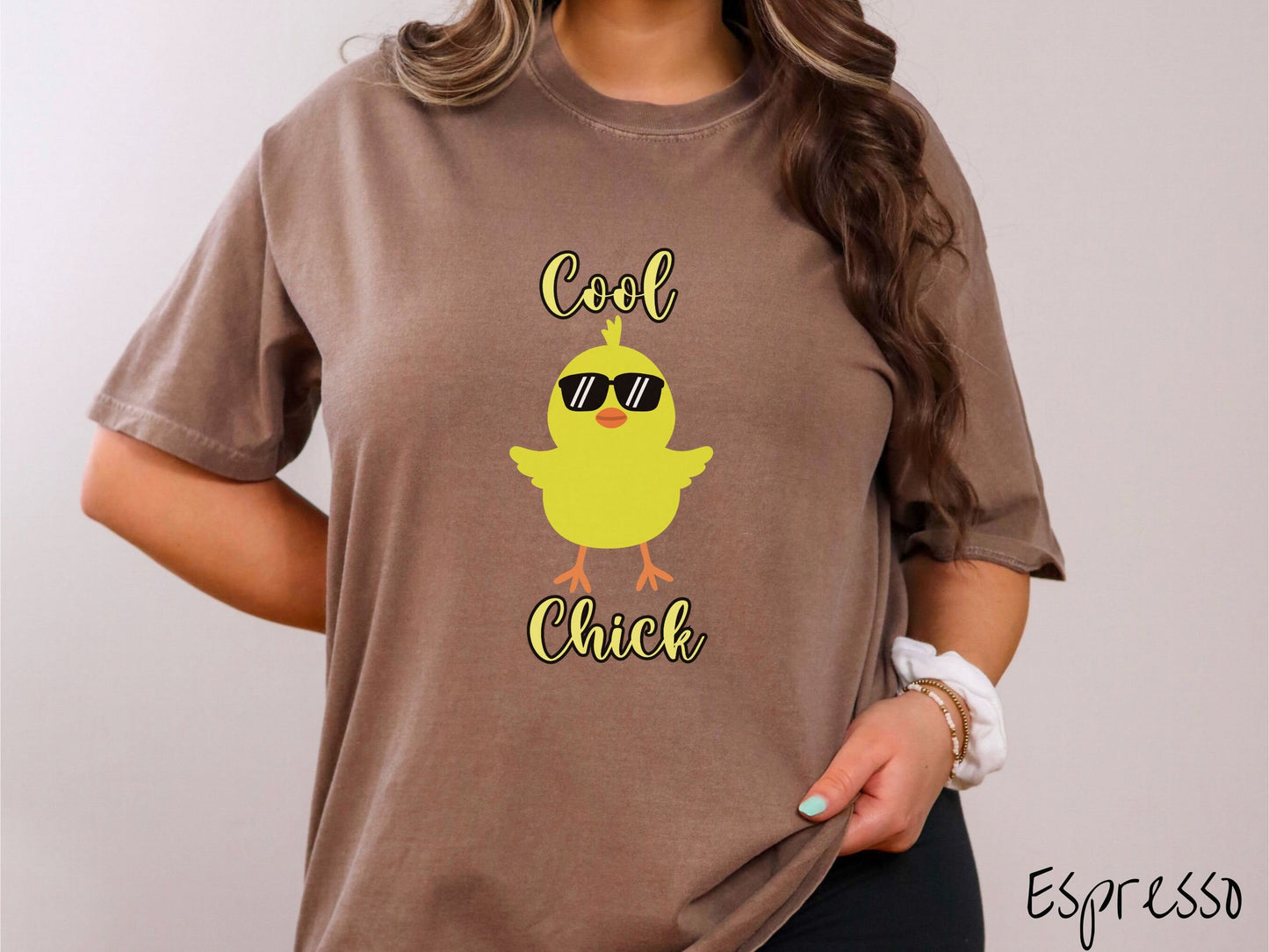 A woman wearing a cute, vintage espresso colored shirt with the yellow text Cool Chick and below is a yellow baby chick with black sunglasses with its wings spread. It has an orange mouth and feet and yellow feathers sticking out the top of its head.
