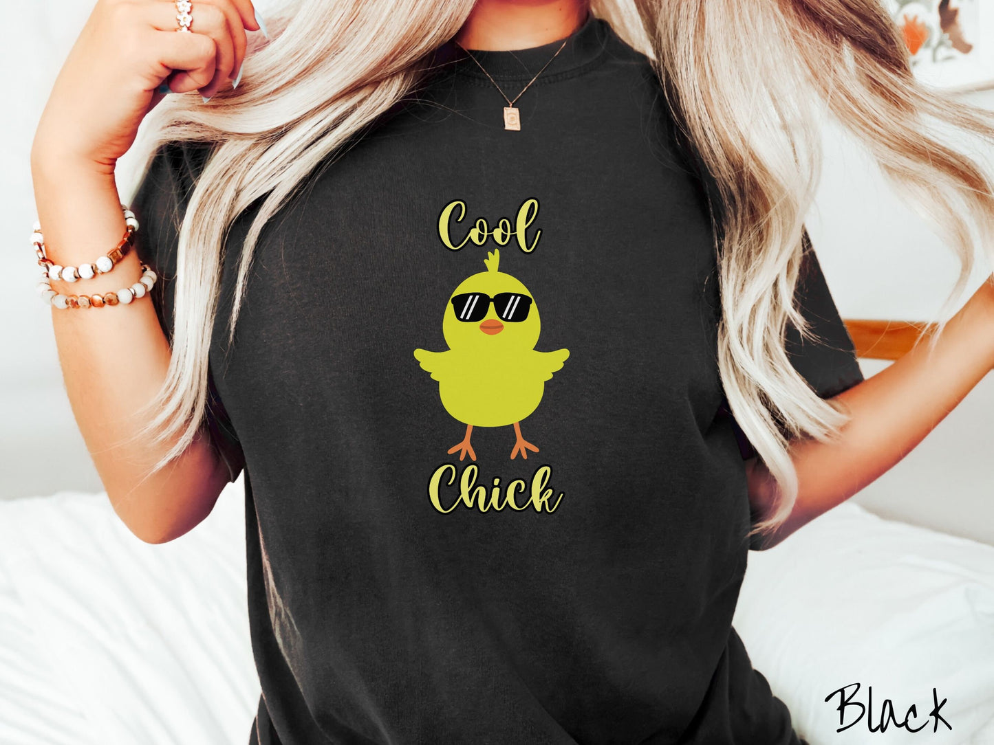 A woman wearing a cute, vintage black colored shirt with the yellow text Cool Chick and below is a yellow baby chick with black sunglasses with its wings spread. It has an orange mouth and feet and yellow feathers sticking out the top of its head.