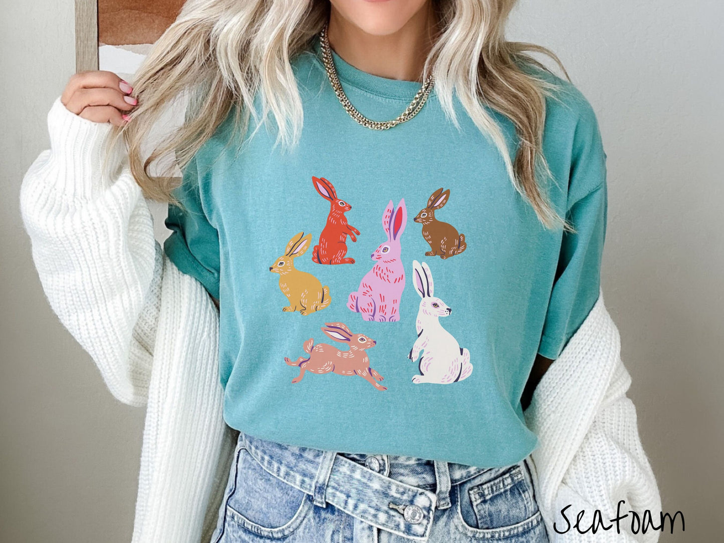 A woman wearing a cute, vintage seafoam colored shirt with six colored bunny rabbits in different stances and postures. One is red, another brown, yellow, pink, light brown, and white.