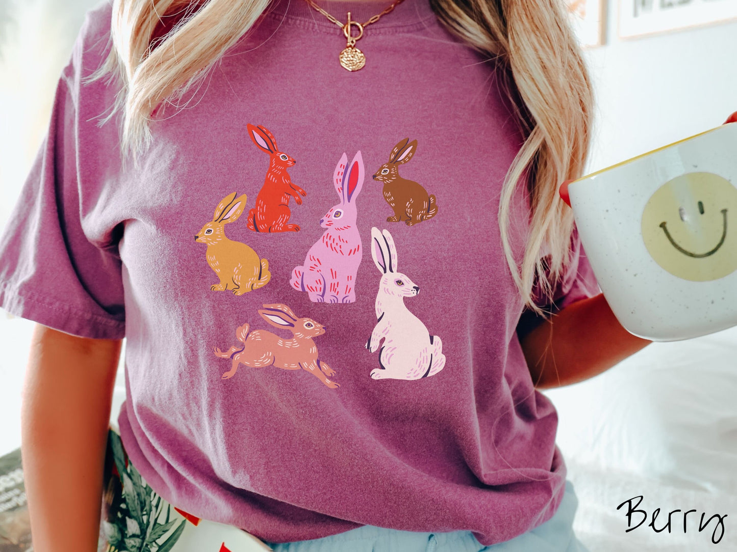 A woman wearing a cute, vintage berry colored shirt with six colored bunny rabbits in different stances and postures. One is red, another brown, yellow, pink, light brown, and white.