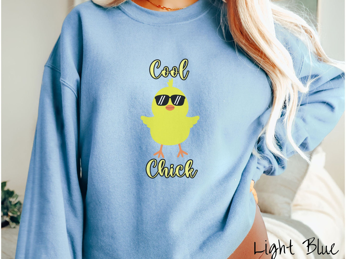 A woman wearing a cute, vintage light blue colored sweatshirt with yellow text Cool Chick and below is a yellow baby chick with black sunglasses with its wings spread. It has an orange mouth and feet and yellow feathers sticking out of its head.
