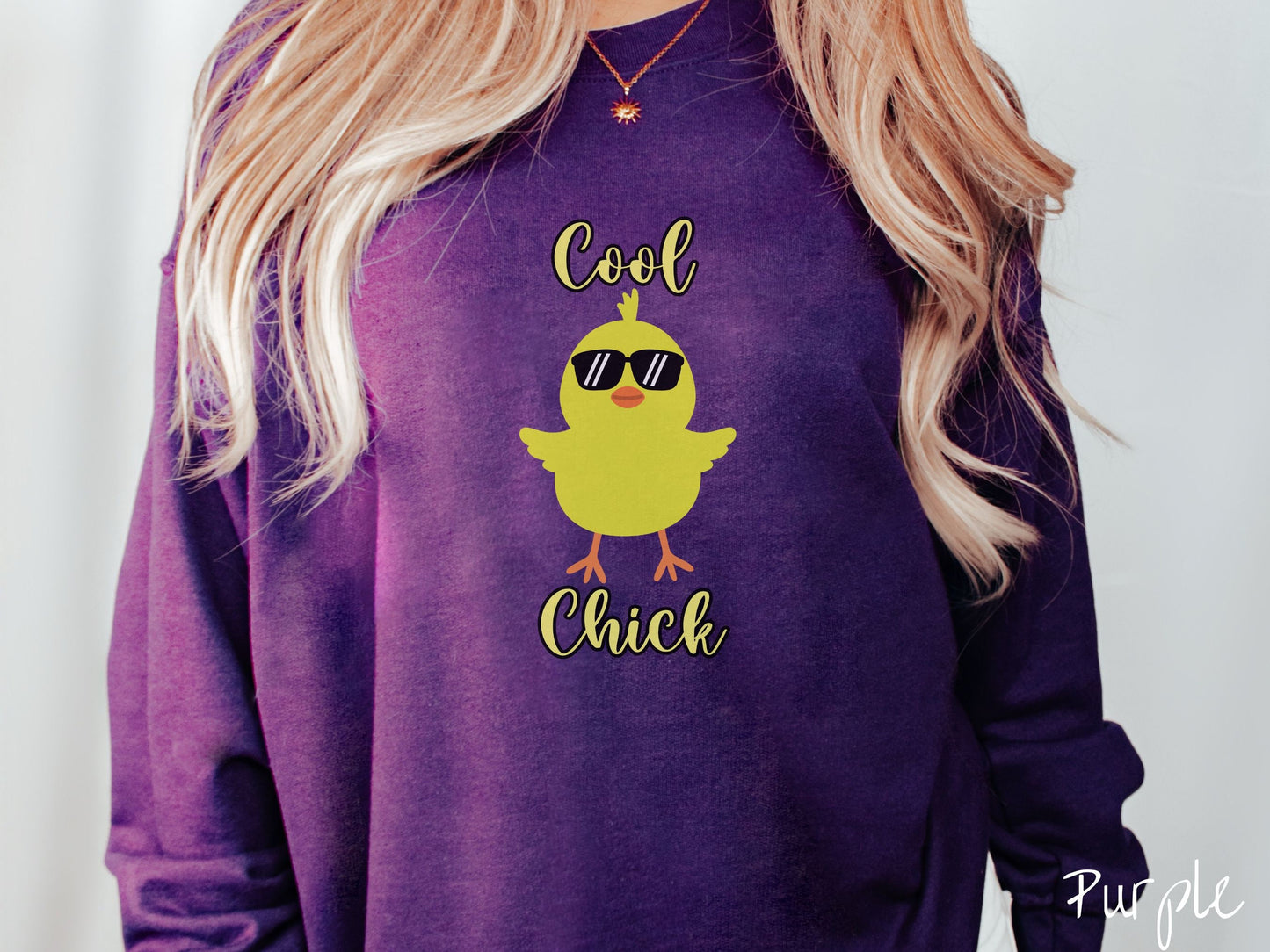 A woman wearing a cute, vintage purple colored sweatshirt with yellow text Cool Chick and below is a yellow baby chick with black sunglasses with its wings spread. It has an orange mouth and feet and yellow feathers sticking out of its head.