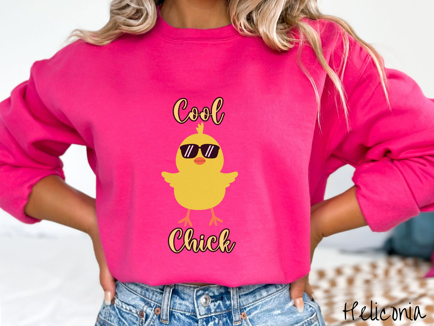 A woman wearing a cute, vintage heliconia colored sweatshirt with yellow text Cool Chick and below is a yellow baby chick with black sunglasses with its wings spread. It has an orange mouth and feet and yellow feathers sticking out of its head.