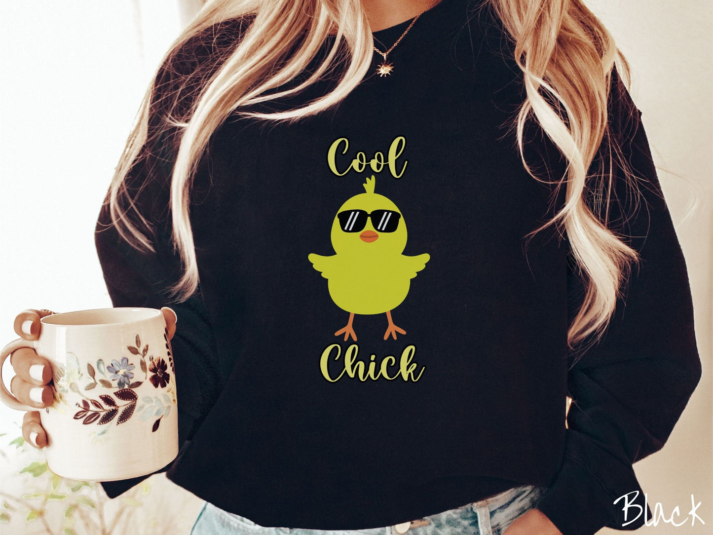 A woman wearing a cute, vintage black colored sweatshirt with yellow text Cool Chick and below is a yellow baby chick with black sunglasses with its wings spread. It has an orange mouth and feet and yellow feathers sticking out the top of its head.