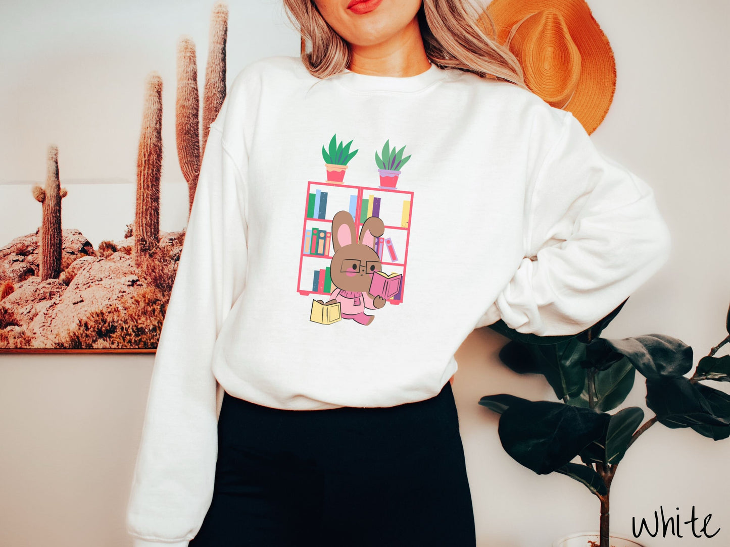 A woman wearing a cute, vintage white colored sweatshirt with a pink-cheeked, brown bunny rabbit wearing glasses reading a pink book in one hand, and a yellow book in the other. In the background is a bookshelf with green plants on top.