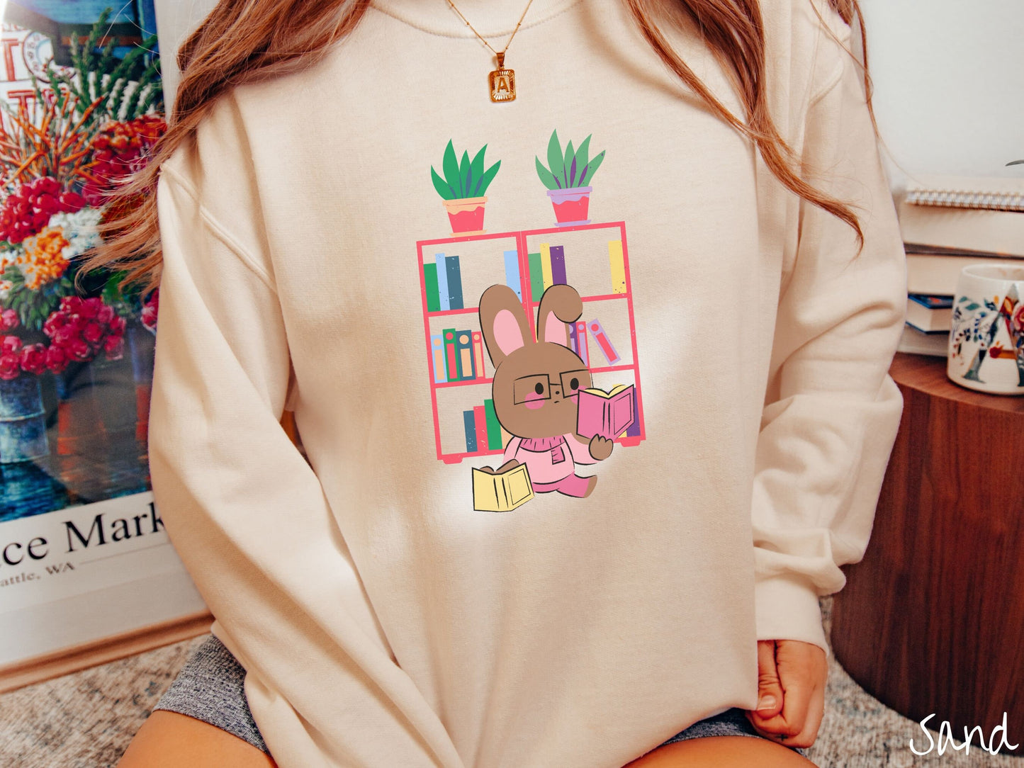 A woman wearing a cute, vintage sand colored sweatshirt with a pink-cheeked, brown bunny rabbit wearing glasses reading a pink book in one hand, and a yellow book in the other. In the background is a bookshelf with green plants on top.