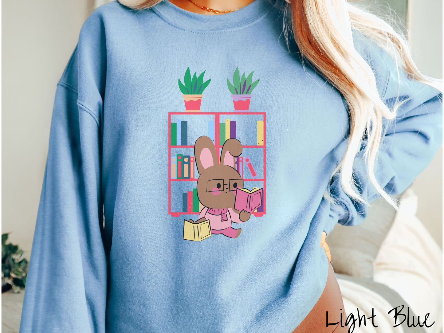 A woman wearing a cute, vintage light blue colored sweatshirt with a pink-cheeked, brown bunny rabbit wearing glasses reading a pink book in one hand, and a yellow book in the other. In the background is a bookshelf with green plants on top.