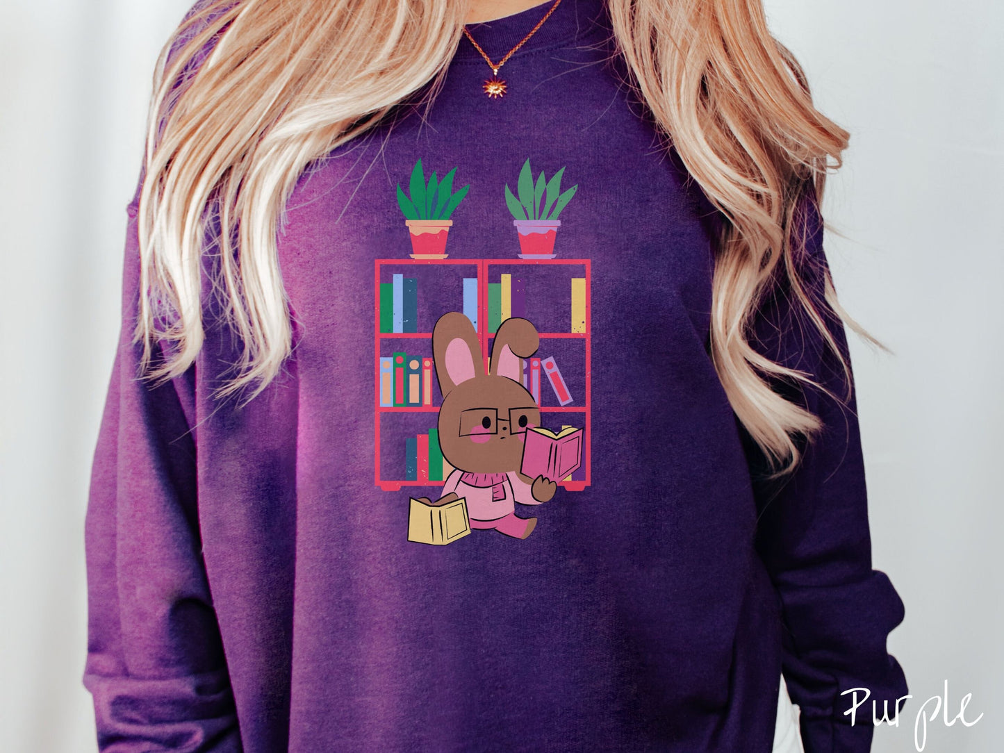 A woman wearing a cute, vintage purple colored sweatshirt with a pink-cheeked, brown bunny rabbit wearing glasses reading a pink book in one hand, and a yellow book in the other. In the background is a bookshelf with green plants on top.