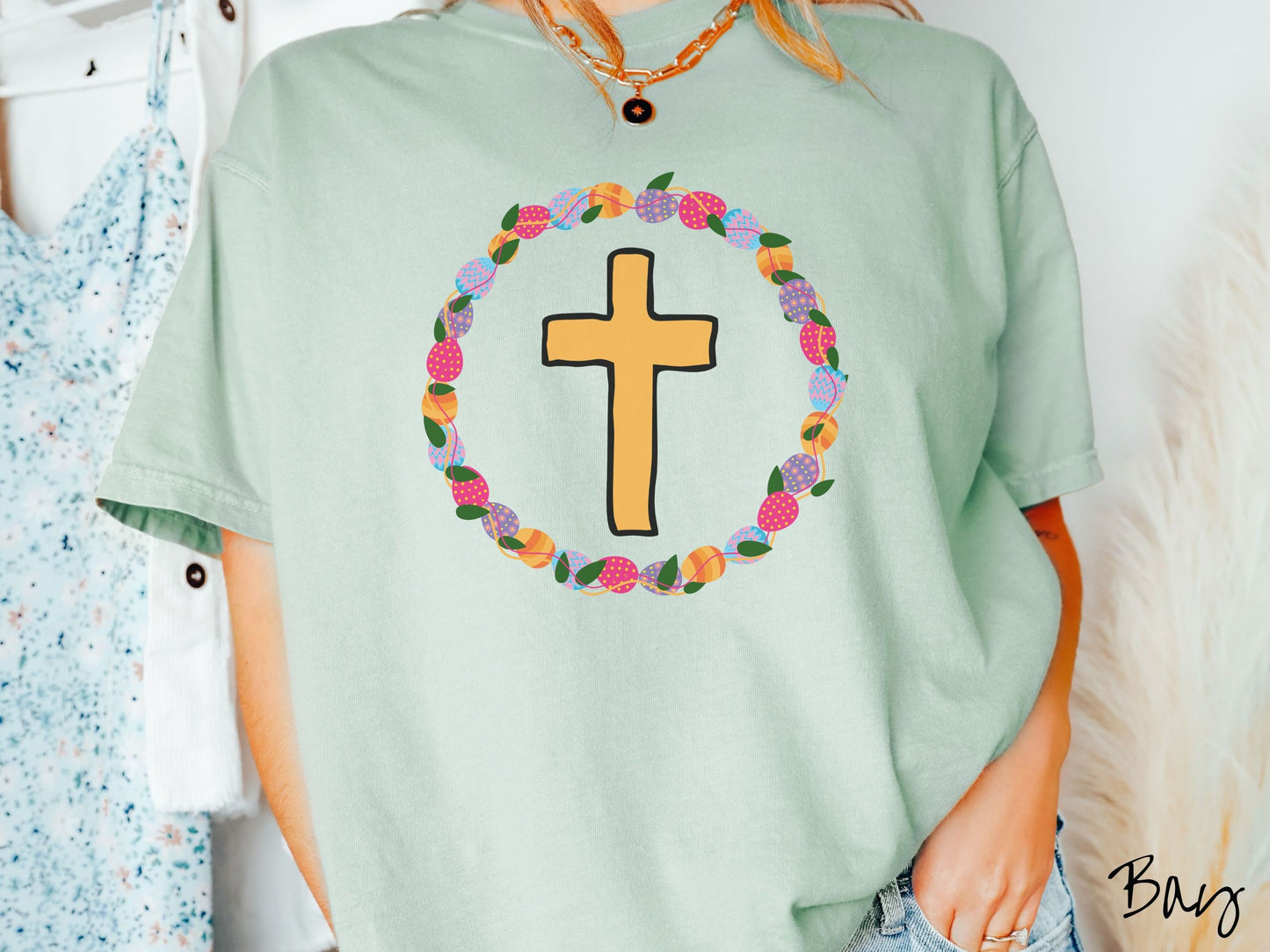 A woman wearing a cute, vintage bay colored shirt with a golden cross with black outline in the center surrounded by a wreath of colorful Easter eggs forming a circle around the cross, intertwined with green leaves.
