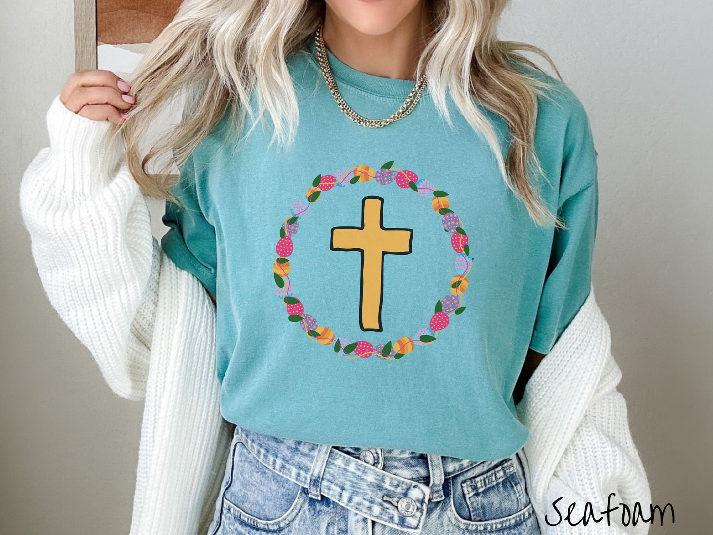 A woman wearing a cute, vintage seafoam colored shirt with a golden cross with black outline in the center surrounded by a wreath of colorful Easter eggs forming a circle around the cross, intertwined with green leaves.
