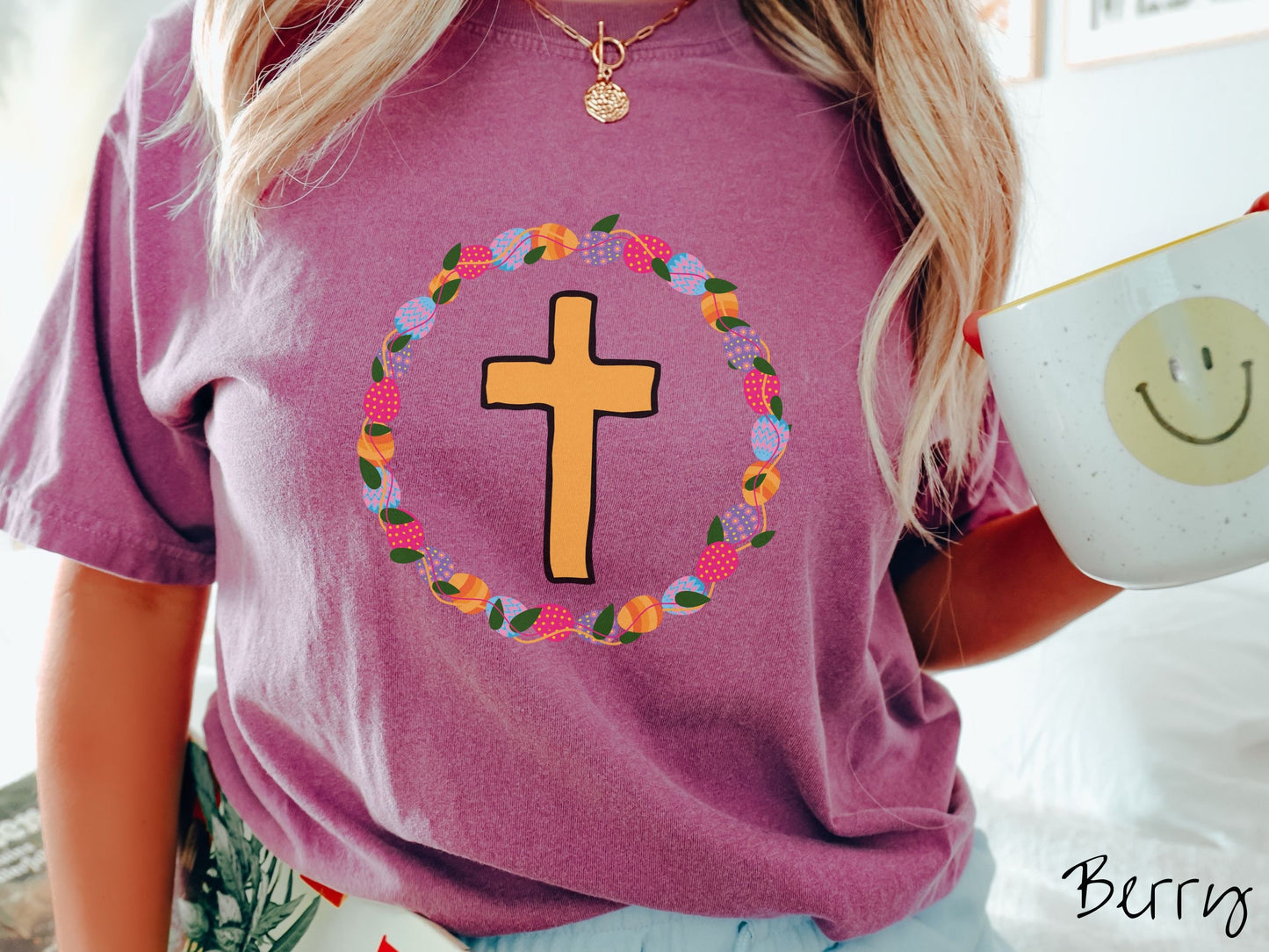 A woman wearing a cute, vintage berry colored shirt with a golden cross with black outline in the center surrounded by a wreath of colorful Easter eggs forming a circle around the cross, intertwined with green leaves.