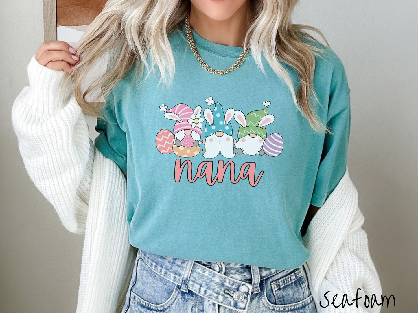 A woman wearing a cute, vintage seafoam colored shirt with text Nana below three gnomes with rabbit ears sitting next to each other. The left one is wearing pink, the middle blue, and right in green, with colorful easter eggs scattered about.
