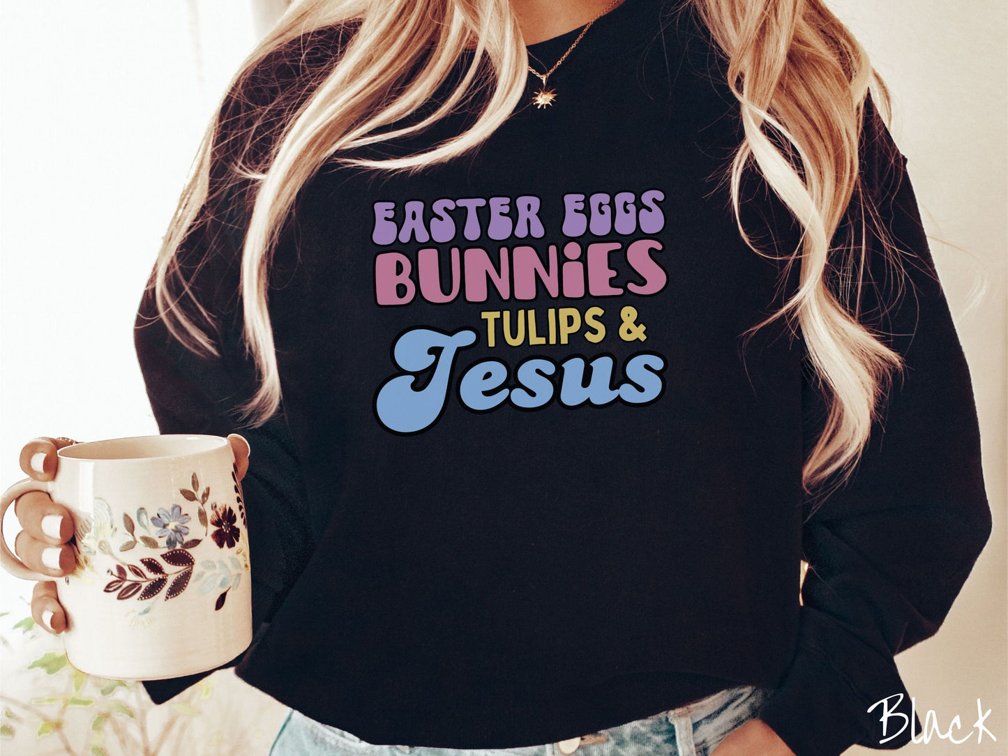 A woman wearing a cute, vintage black colored sweatshirt with text Easter Eggs, Bunnies, Tulips, & Jesus in purple, pink, yellow, and blue font.