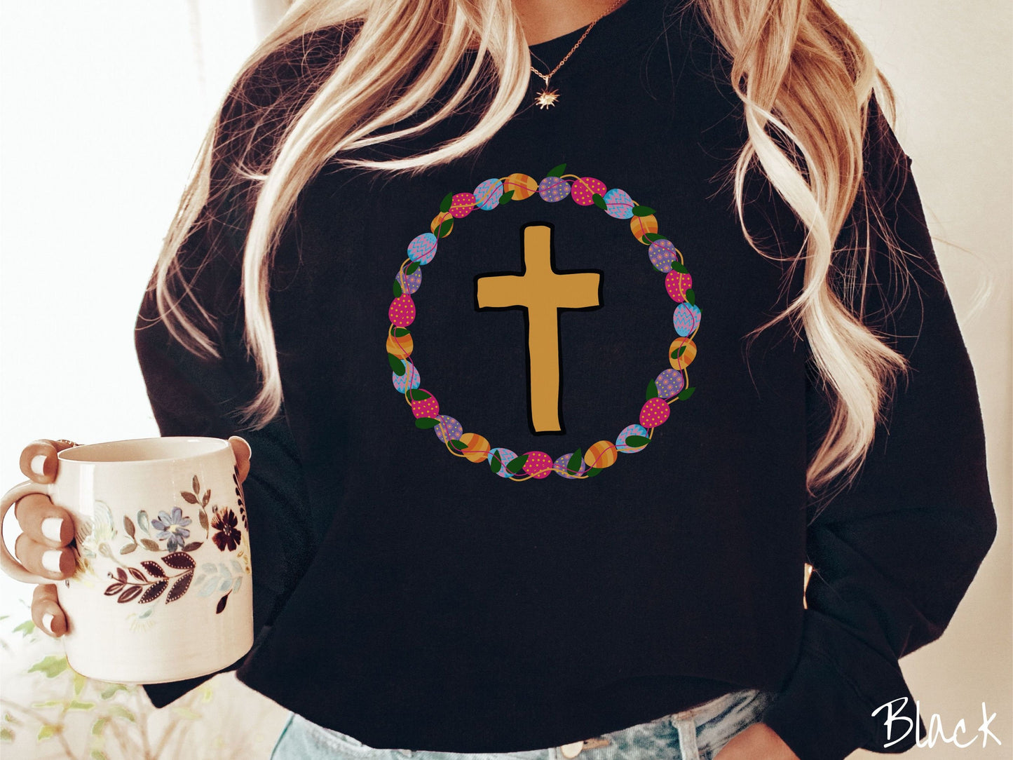 A woman wearing a cute, vintage black colored sweatshirt with a golden cross with black outline in the center surrounded by a wreath of colorful Easter eggs forming a circle around the cross, intertwined with green leaves.