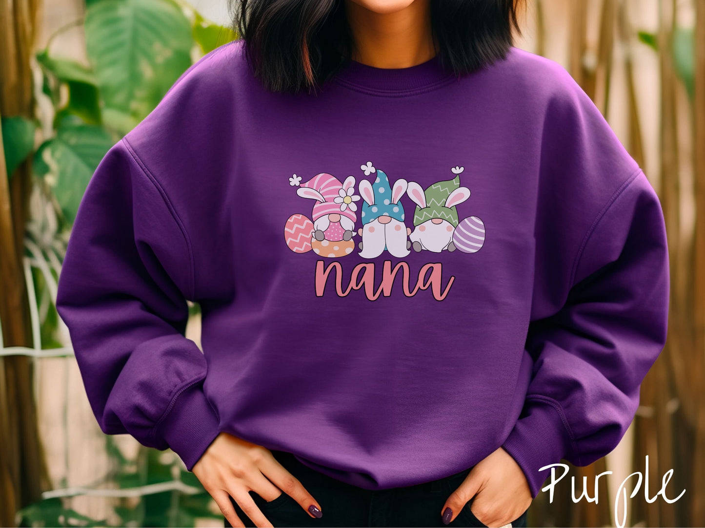 A woman wearing a cute, vintage purple colored sweatshirt with the text Nana below three gnomes with rabbit ears sitting next to each other. The left one is wearing pink, the middle blue, and right in green, with colorful easter eggs scattered about.
