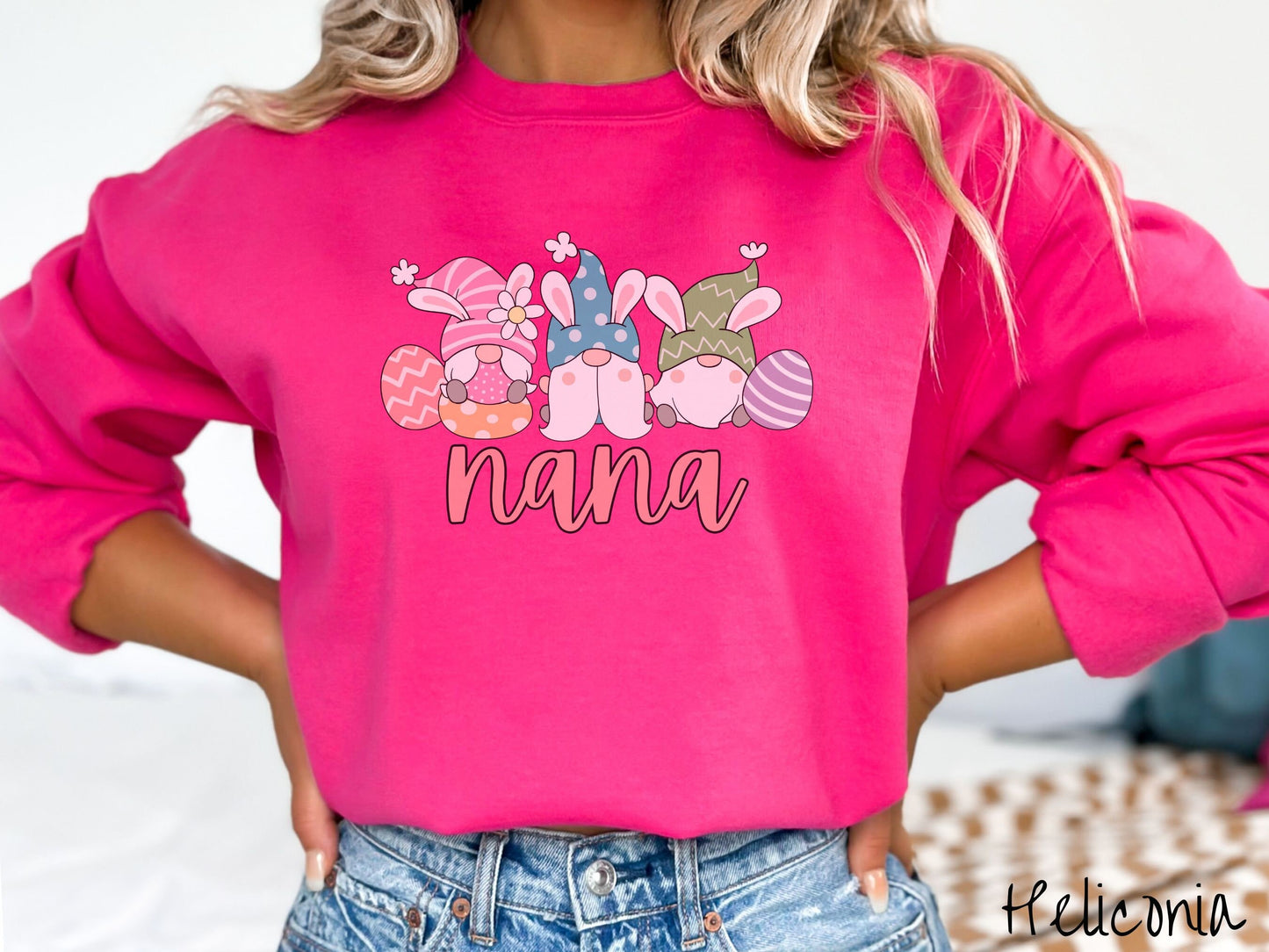 A woman wearing a cute, vintage heliconia colored sweatshirt with text Nana below three gnomes with rabbit ears sitting next to each other. The left one is wearing pink, the middle blue, and right in green, with colorful easter eggs scattered about.