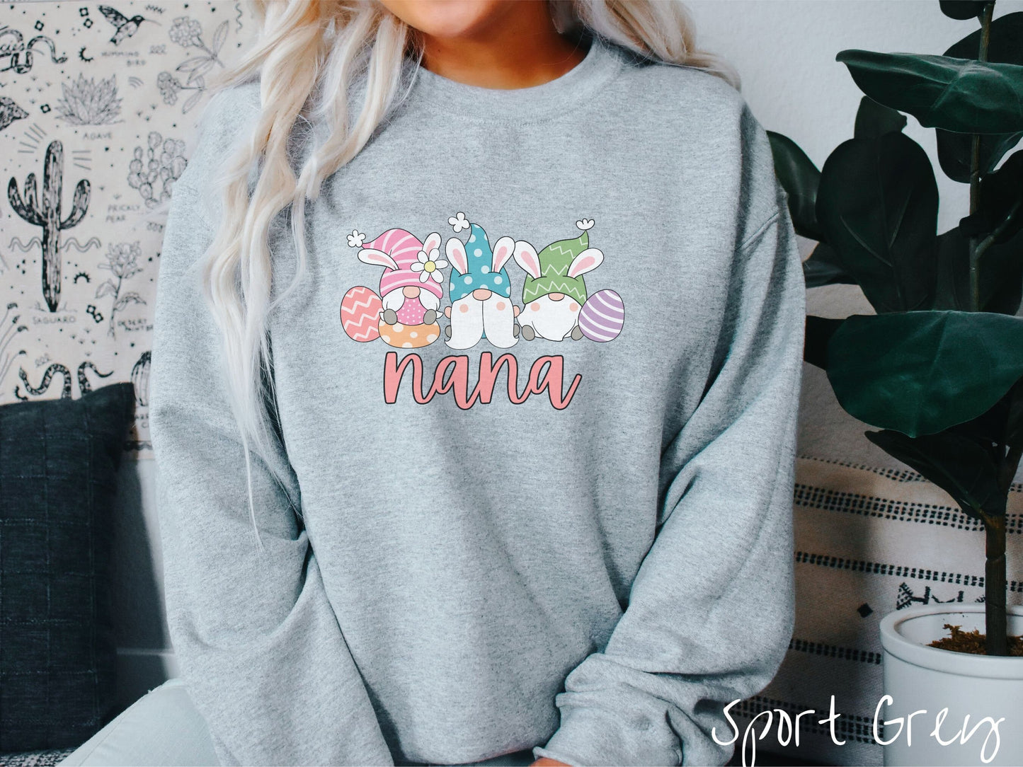 A woman wearing a cute, vintage sport grey colored sweatshirt with text Nana below three gnomes with rabbit ears sitting next to each other. The left one is wearing pink, the middle blue, and right in green, with colorful easter eggs scattered about.