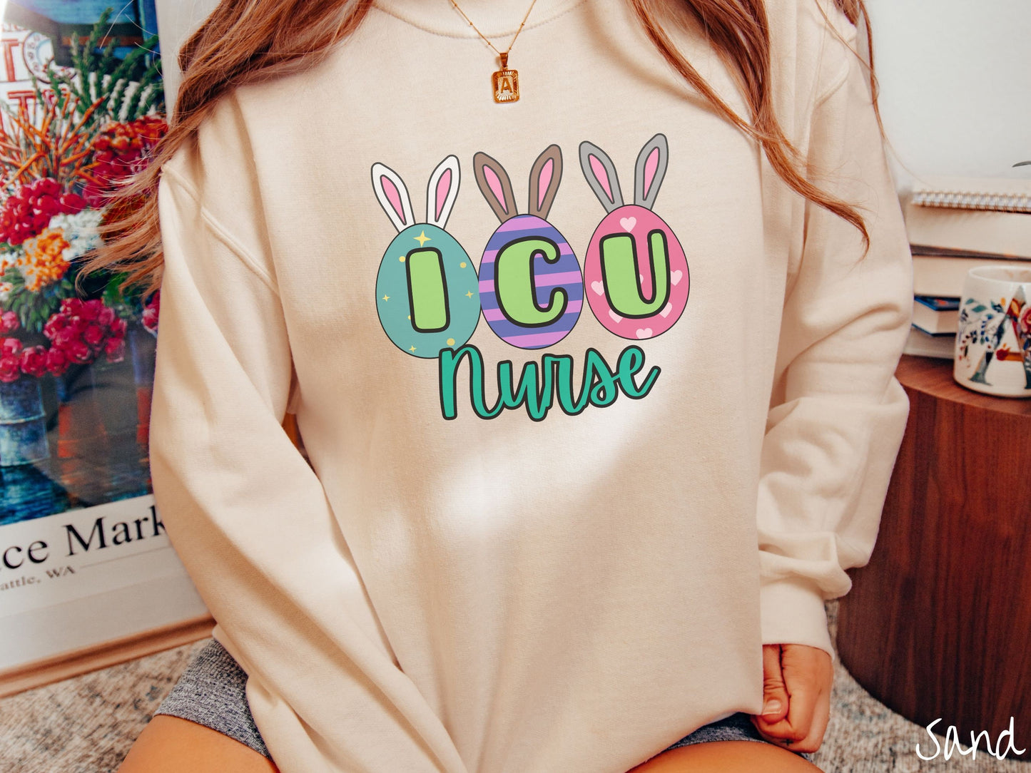 A woman wearing a cute, vintage sand colored sweatshirt with three colorful easter eggs next to each other with the letters I, C, and U inside. Each egg has a pair of bunny rabbit ears, one is white, the middle brown, and the left gray.