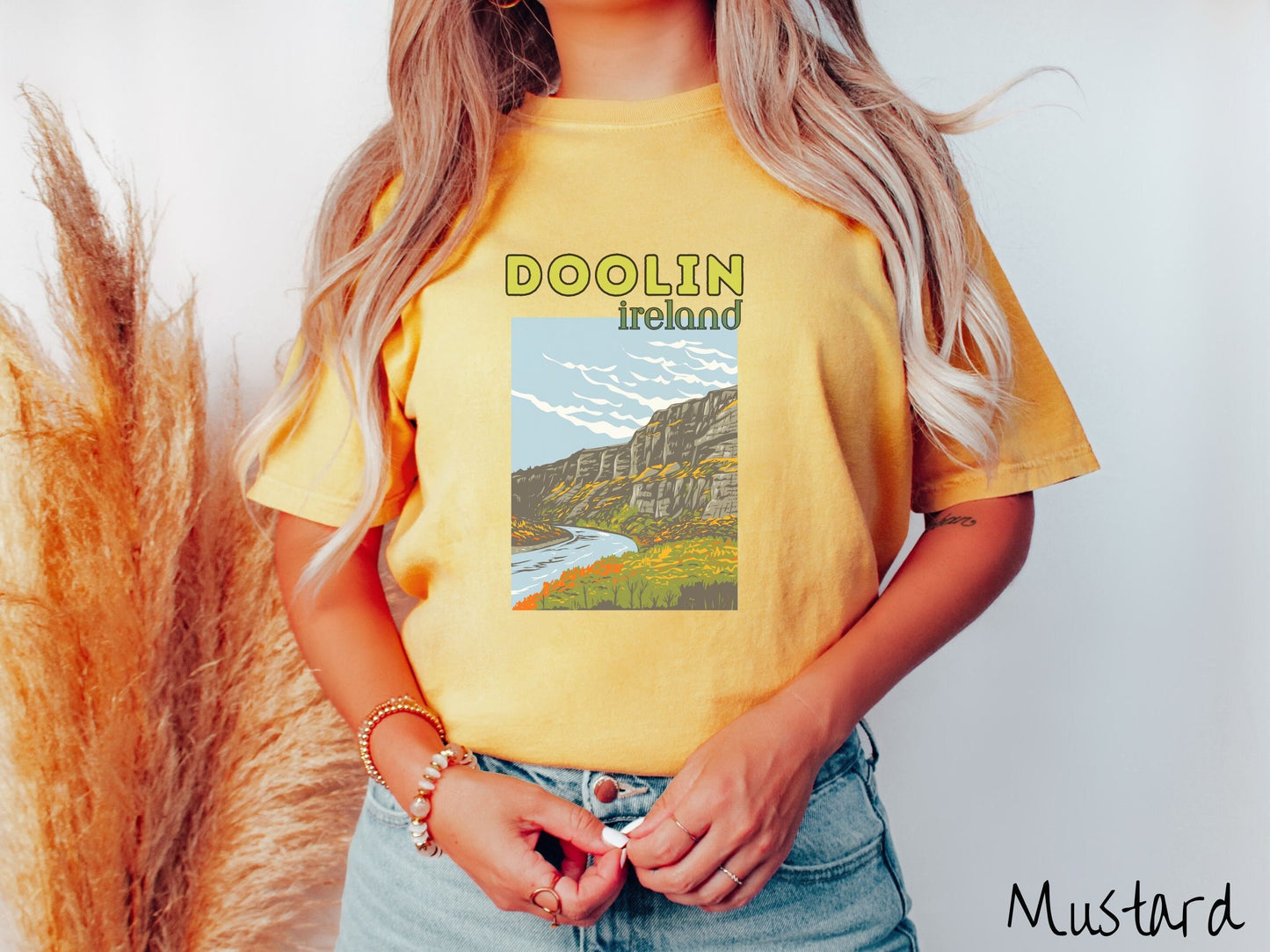 A woman wearing a vintage, mustard colored shirt with the text Doolin Ireland in yellow and green font, respectively, with a picture of an Ireland landscape below, steep cliffs overlooking a rushing river and green meadows in the Irish highlands.