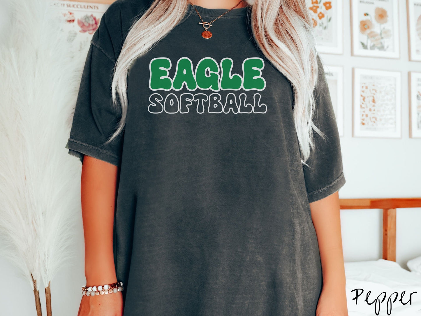 A woman wearing a cute, vintage pepper colored Comfort Colors shirt with the word Eagle across the front in green, uppercase font. Below that is the word Softball in black, uppercase font.