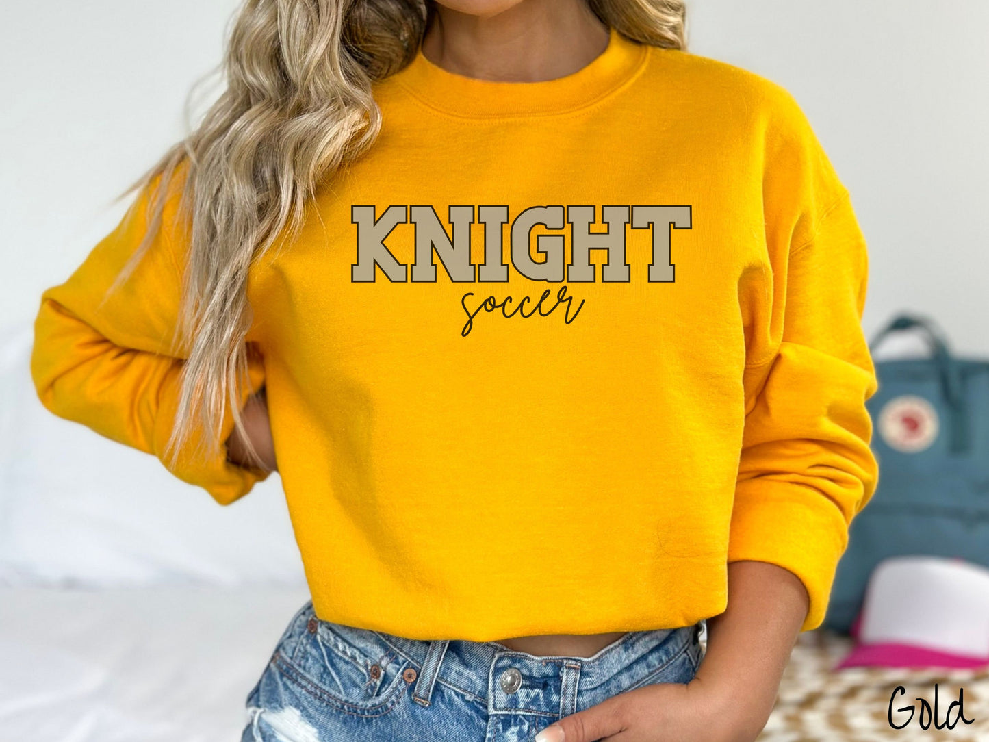 A woman wearing a cute, vintage gold colored sweatshirt with the word Knight in uppercase, gray font across the front and the word soccer below that in black, undercase cursive writing.